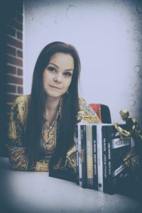 Vera Miettinen istuu pöydän ääressä ja katsoo kameraan, etualalla on hänen kustantamiaan kirjojaan.