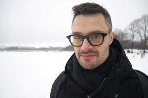 Silmälasipäinen Tommi Peltomäki seisoo lumisen kentän reunalla ja katsoo kameraan