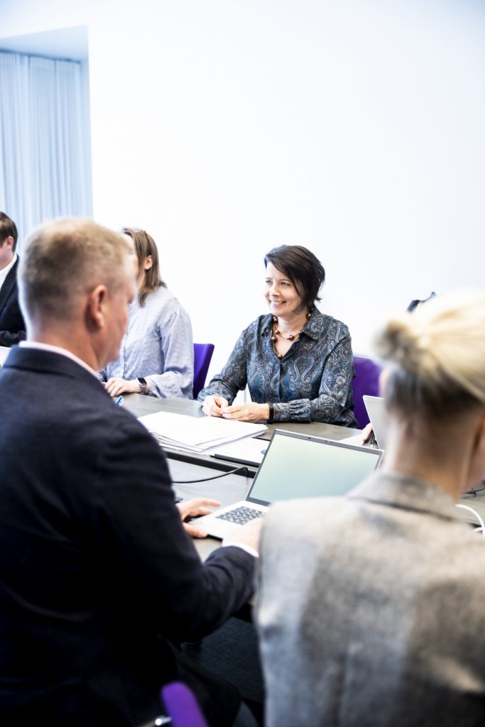 Hanne Aho hymyilee kokouspöydässä palaverin keskellä.