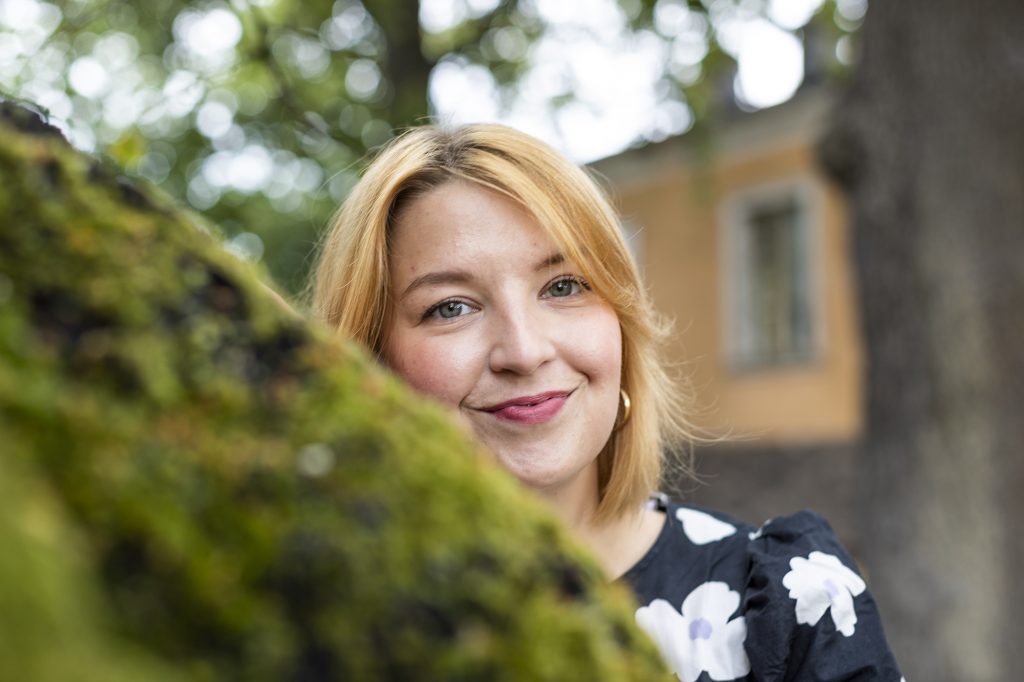 Alma Onali katsoo hymyillen kameraan puun rungon takaa.
