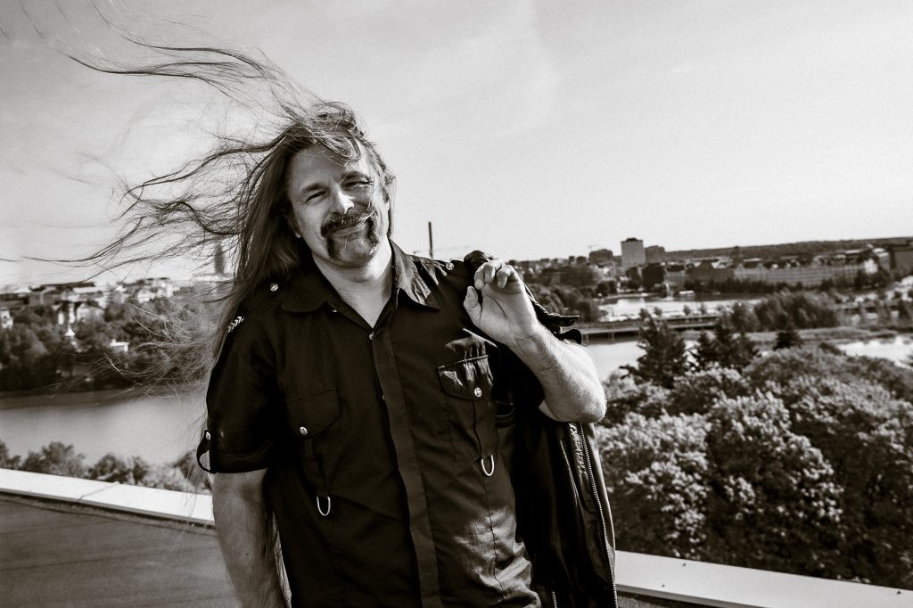 Manu Marttinen seisoo talon katolla rotsi olalla. Tuuli pöllyttää pitkiä rock-uskottaviahiuksia.