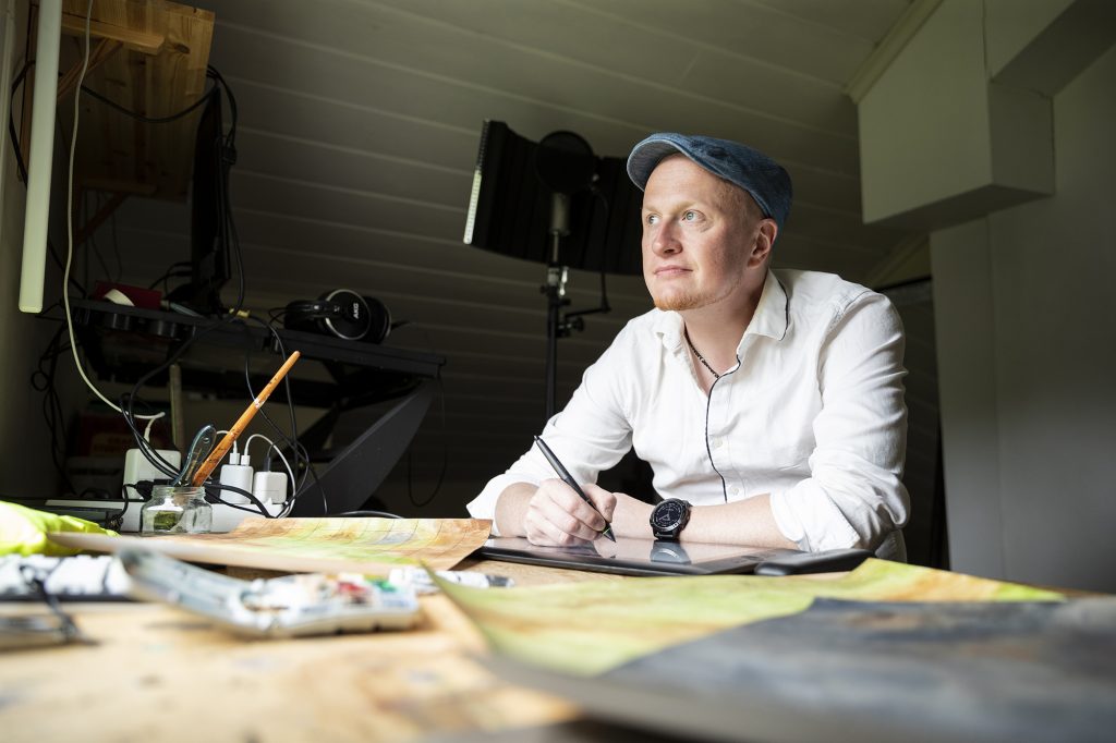 Sebastian Dahlström istuu kotitoimistossaan työpöytänsä ääressä. Tekeillä on jokin kuvitustyö, hän katselee mietteliäänä ulos ikkunasta kynä kädessä, päivänvalo valaisee kasvoja.