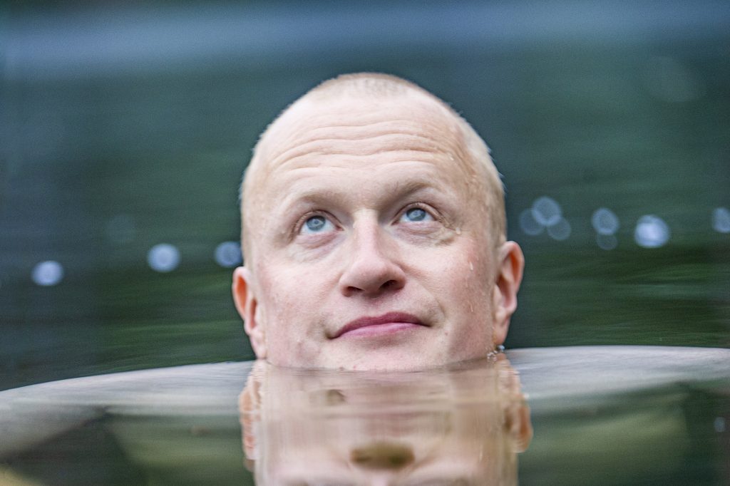 Sebastian Dahlström lammessa. Lähikuvassa hänen vaaleat kasvonsa pistävät esiin tummasta vedestä, silmät katsovat yläilmoihin. Kuvassa on läsnä rauha.