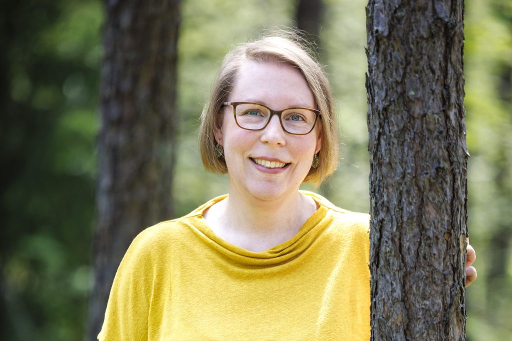 Liina Kjellberg seisoo männynrunkojen välissä T-paidassaan ja hymyilee kameralle kesäisessä metsässä.