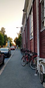 Katukuvassa oikealla puinen työväentalon seinä, jota vastaan nojaa polkupyöriä. Kadulla on rauhallista, muutamia autoja parkissa, ja kaukaisuudessa pyöräiliä. Aurinko laskee ja ilta Kinojuhlilla alkaa.