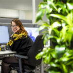 Satu Helin kurkistaa olkansa yli näytön äärestä työpisteeltään. Kuvan vasemmassa laidassa on viherkasveja, joiden yli hänet on kuvattu.